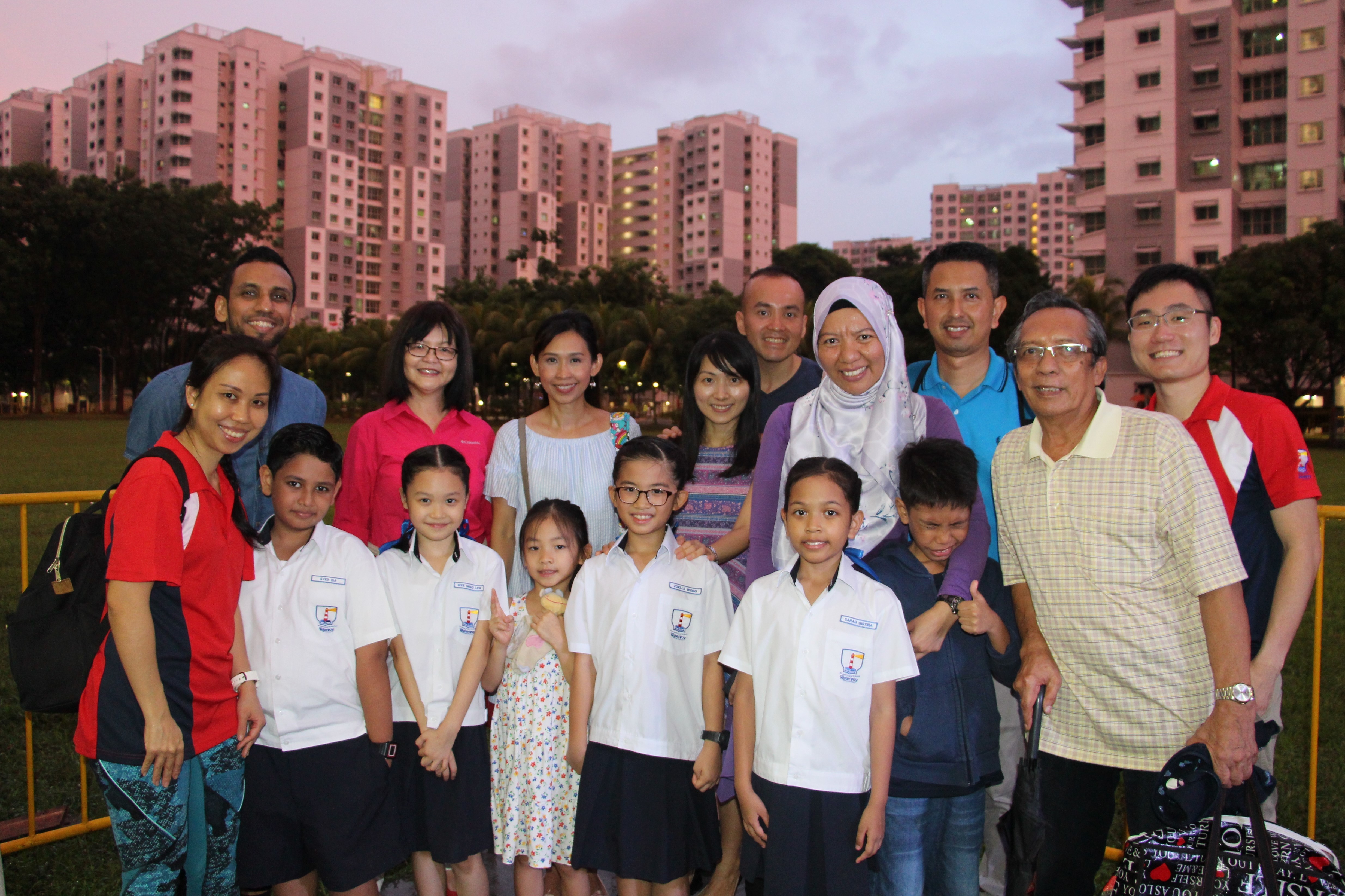 A group of adults and children in school uniforms pose outdoors at dusk with tall pink residential buildings behind them.
